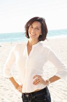 A brunette woman on a beach wearing a thin white blouse and jeans with her hands on her hips