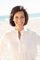 A brunette woman on a beach wearing a thin white blouse