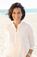 A brunette woman on a beach wearing a thin white blouse