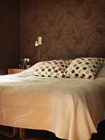 Kissen mit Punktemuster und helle Tagesdecke auf Doppelbett, vor tapezierter Wand mit floralem Muster auf braunem Untergrund