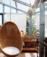 Gemütlicher Hängekorbsessel mit weißem Sitzpolster und rustikaler Hobelbank im Glashaus