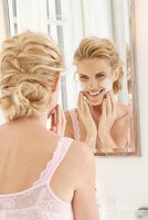 Blonde Frau vor dem Spiegel trägt Reinigungsmilch im Gesicht auf