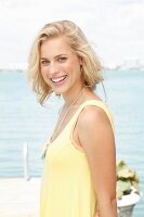A blonde woman by the sea wearing a sleeveless, pastel yellow top