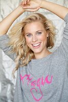 A blonde woman holding her arms above her head wearing a grey jumper with the word 'Magic' in pink