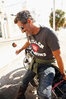 Mann in Jeans, T-Shirt und Sonnenbrille sitzt seitlich auf Motorrad