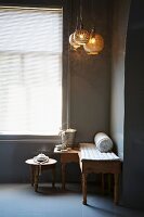 Carved bench and side table below pendant lamps next to window with closed blinds in corner of grey-painted room