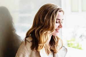 Junge, blonde Frau mit heller Jacke sitzt am Fenster