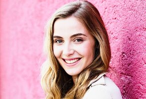 A portrait of a dark blonde woman against a pink wall