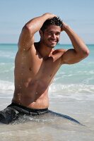 A young, topless man wearing bathing shorts keeling on the beach in the water