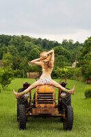 Frau in Unterwäsche mit Strohhut sitzt rücklinks auf altem Traktor