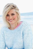 A blonde woman on a beach wearing a pink t-shirt and a fluffy jumper