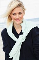 A blonde woman on a beach wearing a dark woollen jumper with a white jumper over her shoulders