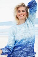A blonde woman on a beach wearing a blue angora jumper with her arms above her head