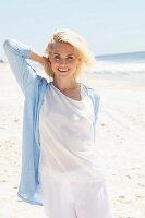 A blonde woman on a beach wearing a white shirts, trousers and a light blue cardigan