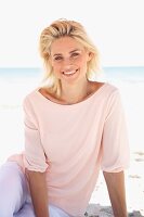 Lächelnde blonde Frau in pastellfarbenem Shirt und Hose sitzt am Strand