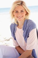 Lächelnde blonde Frau in pastellfarbenem Shirt, Pulli und Hose sitzt am Strand
