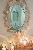 Romantic, ornate oval mirror reflecting bathroom above arrangement of roses and tealight holders