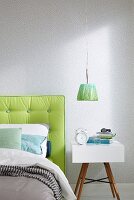 A modern bedside table next to a bed with a lime green padded headboard against a wall with a light-coloured liberty pattern