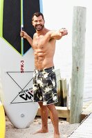 A dark-haired man on a beach wearing bathing shorts with a surfboard