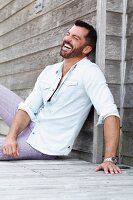 A dark-haired man with a beard wearing a white shirt and checked trousers sitting on the floor laughing