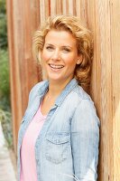 A blonde woman wearing a denim shirt leaning on a fence