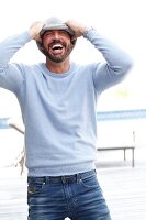 Mann mit Vollbart in hellblauem Pullover und Jeans macht Spaß mit Strickmütze