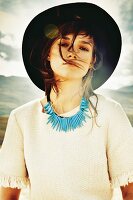 A brunette woman wearing a hat, I white blouse with tassels and blue necklace