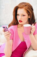 Rothaarige Frau in pinkfarbener Bekleidung mit Kaffeebecher und Smartphone