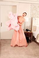 A young woman in a hotel room wearing an evening dress and holding pink plush elephant