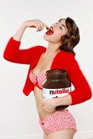 Junge Frau in Unterwäsche und Strickjäckchen nascht aus großem Glas Nutella