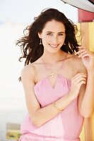 A young brunette woman wearing a pastel pink summer dress leaning against a wooden wall