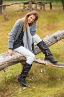 Junge Frau in grauer Strickkleidung und Lederstiefeln sitzt auf Baumstamm