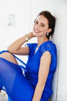 A brunette woman wearing a blue dress sitting against a wooden wall