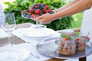 Glass dish of fresh berries held in hands and preserving jars on tray on garden table