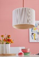 A pendent lamp with light fabric over bent wooden veneer over a table laid with buttercups against a pink wall