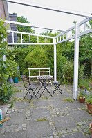 Garden chairs and a table on a terrace with a white painted pergola frame and paving slabs