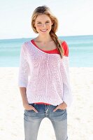 A young woman on a beach wearing a red top, a white open-work jumper and jeans
