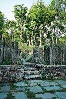 A view of steps and an open gate with a stave fence on a natural stone wall with a spacious garden and high trees in the background