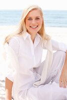 Junge blonde Frau in weisser Bluse und Hose sitzt am Strand