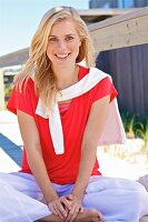 A young blonde woman sitting on a wooden jetty wearing a red top and white trouser