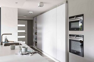 Detail of a kitchen counter opposite a modern, built-in kitchen cupboard in white in an open-plan kitchen