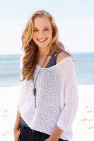 A young blonde woman on a beach wearing a tank top and a net jumper