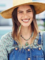 A woman wearing a patterned blouse, a denim pinafore dress and a straw hat with a blade of grass in her mouth