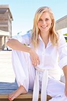 A young blonde woman sitting on a wooden jetty wearing a white blouse and trousers