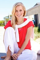 A young blonde woman sitting on a wooden jetty wearing a red top and white trouser