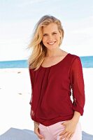 A young blonde woman on a beach wearing a bordeaux red top and light trousers