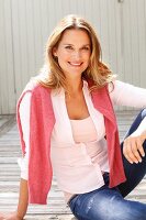 A blonde woman wearing a pink top, blouse and jeans with a pink jumper over her shoulders