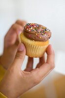Hands holding a cupcake topped with chocolate cream and colourfu hundreds and thousands