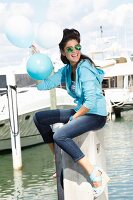 Brünette Frau in Jeans, Ringelshirt, Hoodie, Wedges und Sonnenbrille sitzt auf Betonpfeiler am Hafen mit Luftballons in der Hand
