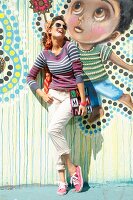 A brunette lady wearing beige-coloured chinos, a striped jumper, sunglasses, a hairband and sneakers leaning against a colourful exterior wall